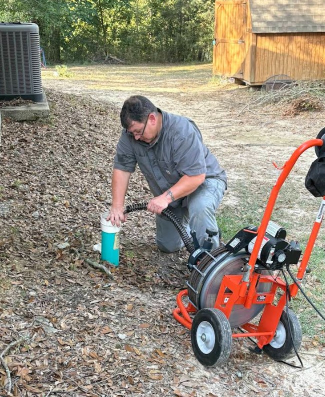 Man kneels in a yard, using a plumbing machine to clear a drain pipe. The setting is outdoors, with leaves scattered around and a shed in the background.