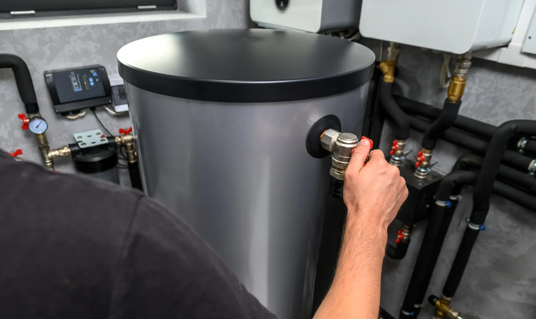 A person adjusts a valve on a large, metallic water heater in a utility room. The space contains various pipes, gauges, and control panels.