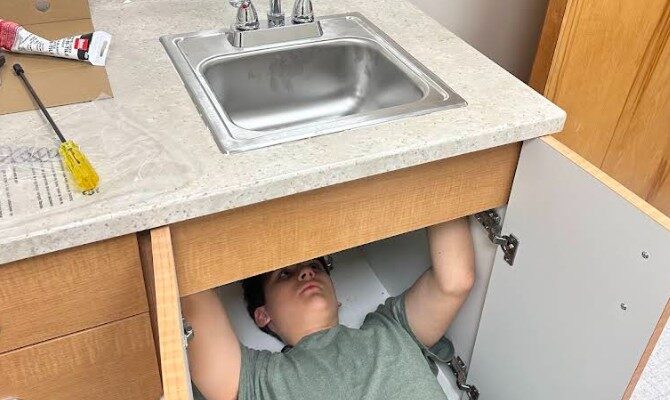 A person in a green shirt lies under a kitchen sink, with cabinet doors open. Tools are nearby, suggesting a plumbing task. The scene feels focused and practical.
