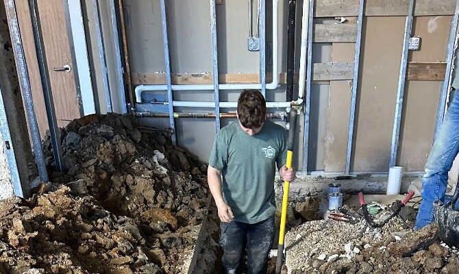 Two workers in green shirts and jeans are digging trenches inside a partially constructed building. One holds a yellow shovel. Soil piles and exposed pipes visible.